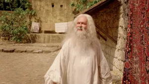 Een foto waarin God wordt uitgebeeld in een wit gewaad met een lange baard. God loopt naast een woning, op weg om een uitverkoren volk te kiezen. Naast god hangt een Perzisch tapijt aan de muur. Achter god hangen doeken en tapijten op een waslijn. 