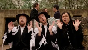 Een foto van twee mannen en één vrouw in traditioneel zwarte en witte Joodse kleding. Ze houden hun handen in de lucht in een zwaaiende beweging. Ze zitten voor een lage muur en achter de muur staan drie andere Joodse mensen te praten.