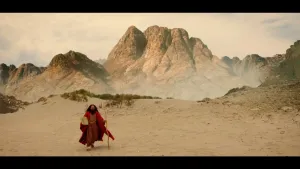 Een foto van Mosje (Mozes) die door een zandvlakte loopt met op de achtergrond bergen. Mosje (Mozes) is op de terugtocht van de berg Sinaï. Hij draagt de tien geboden onder zijn rechterarm en een houten tak als wandelstok in zijn linkerhand.  