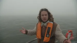 Een foto van presentator Pascal die op een boot staat en voor de camera iets uitlegt. Op de achtergrond is er mist en water te zien.
