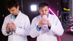 Een foto van presentatoren Jurre en Sahil die allebei een ballon aan het opblazen zijn in een doorzichtige plastic fles.