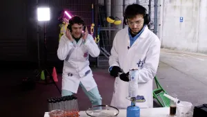 Een foto van presentatoren Jurre en Sahil die, met een veiligheidsbril en oorbeschermers op, een experiment uitvoeren. Sahil houdt met een tang een limonadefles bij een gasbrander. Jurre staat erachter met zijn handen naast zijn gezicht.