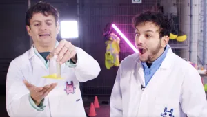 Een foto van presentatoren Jurre en Sahil. Jurre heeft links een glazen maatbeker vast waar een geel post-it briefje aan vastgekleeft zit. Sahil kijkt rechts toe met zijn mond open en een verbaasde blik..