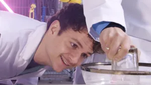 Een foto van presentator Jurre, die met zijn gezicht dichtbij een kom met water staat. Daarnaast is de arm van presentator Sahil te zien, die een glas vastheeft dat omgekeerd (met de bodem omhoog) in de kom met water staat.