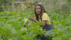 Een foto van presentator Eva die tussen de groenteplanten zit. Ze houdt er een vast in haar hand terwijl ze er iets over uitlegt in de camera.