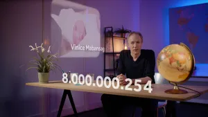 Een foto van de presentator die achter een tafel zit. Achter hem is een foto van een baby met de naam Vinice Mabansag. Voor hem staat op de foto het getal 8.000.000.254.