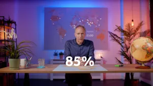 Een foto van de presentator die achter een tafel zit. Achter hem hangt een wereldkaart met witte stippen, en de tekst "Megasteden 2100". Voor hem staat "85%".