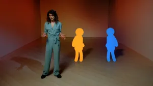 Een foto van een presentator met daarachter twee getekende figuren van een mens die naar elkaar toe gericht staan en hun schaduwen. Eén van de menselijke figuren is volledig oranje en één volledig blauw van kleur.