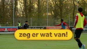 Een foto van een voetbalveld met daarop vier voetballers. Op de voorgrond is de geel omrande tekst "zeg er wat van!" zichtbaar.