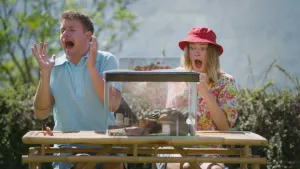 Een foto van presentatoren Jurre en Emma die achter een tafel zitten waar een bak op staat waar je doorheen kan kijken. In de bak liggen wat stenen waar een slang bovenop ligt.