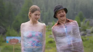 Een foto van presentatoren Emma en Jurrie die staan terwijl ze gewikkeld zijn in plastic waar je doorheen kan kijken. Op de achtergrond is een bos te zien.