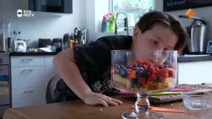 Een foto van een kind in een keuken. Hij staat voorover gebogen achter een tafel met een glazen schaal. In de schaal liggen stukken cake en verschillend fruit.