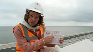 Een foto van presentator Pascal die op de afsluitdijk zit met een helm op waar 'nummer 1' op staat. Hij heeft een kaart van Nederland in zijn handen en laat die zien in de camera terwijl hij op de kaart wijst naar het IJsselmeer..