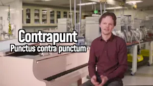 Een foto van een presentator achter een witte piano. Naast de presentator staat aan de linkerkant de tekst: "Contrapunt Punctus contra punctum". Op de achtergrond is een laboratorium te zien met grote machines op een tafel.