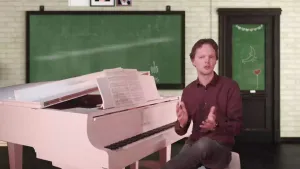 Een foto van een presentator die achter een witte piano zit. Op de achtergrond is een schoolbord zichtbaar met daarop met krijt een witte, van links naar rechts stijgende, lijn getekend.