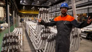 Een foto van presentator die met zijn armen wijd staat. Hij staat in een fabriek met allemaal metalen buizen op de achtergrond. Hij draagt een helm en bril.