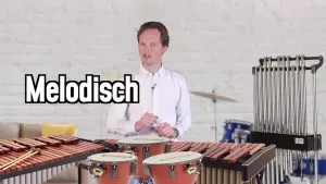 Een foto van een presentator met allemaal muziekinstrumenten die met een slagtechniek bespeeld worden. De instrumenten marimba, xylofoon, buisklokken en pauken zijn voor de presenator te zien op de foto. Op de achtergrond is ook een drumstel te zien.