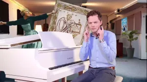 Een foto van een presentator die achter een witte piano zit en naar zijn oren wijst. Op de achtergrond is een foto van de actrice Julie Andrews te zien uit de film 'The Sound Of Music'. Ook is de voorkant van Guido van Arezzo's muzieknotatie zichtbaar.