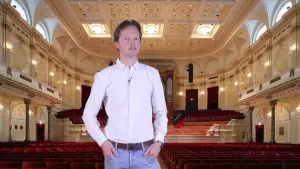 Een foto van een presentator met op de achtergrond een foto van een concertgebouw waarop de zaal met stoelen en een gedeelte van het podium achter de presentator zichtbaar zijn. Het concertgebouw is speciaal geschikt voor het beluisteren van live muziek.