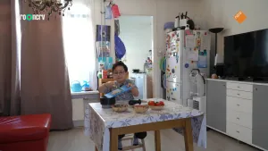 Een foto van een jongetje die in een keuken aan een eettafel zit. Hij schuift wat voedsel met een mes van een snijplank af in een schaal die op tafel staat.