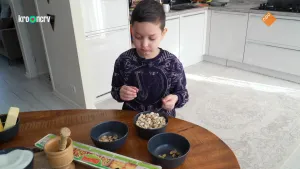 Een foto van een kind in een keuken. Hij staat achter een tafel en kijkt naar wat op tafel staat. Er staan bakjes met nootjes en andere dingen om mee te bakken.