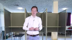 Een foto van een presentator met op de achtergrond stemhokjes in een stemlokaal dat gebruikt worden voor verkiezingen. Verkiezingen worden ook wel stemmingen genoemd.