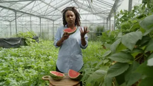 Een foto van presentator Eva in een gebouw van glas met heel veel planten. Ze staat achter een tafeltje. Op het tafeltje en in haar hand zijn stukken watermeloen.