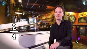 Een foto van een presentator die achter een witte piano zit. Links van de piano is de maatsoort "twee vierde" leesbaar. Achter de presentator is een bar zichtbaar met flessen drank op planken aan de muur. Ook hangt er rechts een dartbord.