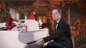 Een foto van een presentator in een zwart pak, die achter een witte Yamaha piano zit. Op de piano staat kerstversiering zoals een knuffel van een rendier, een kerstman, een kerstmuts en een kleine, roodgebladerde kerstboom.