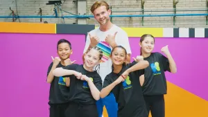 Een foto van danscoach Olivier Feikens die samen met Deliano, Lewis, Nora en Estrella van Kinderen voor Kinderen op de foto staat. Ze gebaren blij met duimen omhoog of vuisten tegen elkaar. De kinderen dragen een Kinderen voor Kinderen t-shirt.