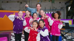 Een foto van een groep kinderen in een grote zaal met kleurrijke jassen in rood, roze of paars met hun naam erop. De kinderen vormen met hun handen allemaal het hartjessymbool. Achter de kinderen staan zangcoach Michiel en een elektrische piano.