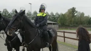 Een foto van twee politiepaarden met op ieder paard een politieman in werkkleding. Op de achtergrond is een rijbak voor de paarden zichtbaar. Rechts op de foto staat de presentator naar de voorste politieman toegekeerd.