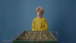 Een foto van een jongen achter het tafelvoetbalspel van Nico. De jongen speelt niet met het tafelvoetbalspel, maar kijkt naar de persoon waar hij tegen praat die niet in beeld is.