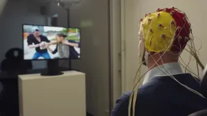 Een foto van een man met een muts op die voor de wetenschap wordt gebruikt. Op de muts zitten veel meters bevestigd waar draadjes uitkomen. De man zit televisie te kijken.
