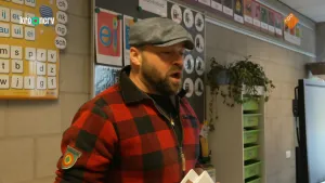 Een foto van presentator Tim die in een klaslokaal staat. Achter hem hangen er borden aan de muur met daarop verschillende letters en woorden.