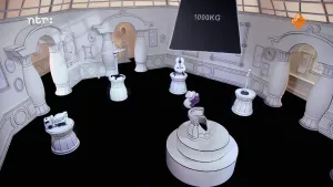 Een foto van een zwart-witte kamer, met in het midden een stoel. Boven de stoel hangt een groot blok met de tekst "1000KG". Achter de stoel staat een paarse pop.