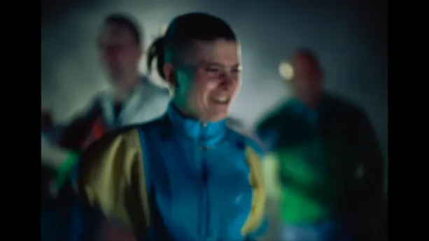 Een onscherpe foto met een gabber in een trainingspak die lachend op de voorgrond staat. Op de achtergrond zijn nog onscherper twee gabbers te zien die aan het dansen zijn.