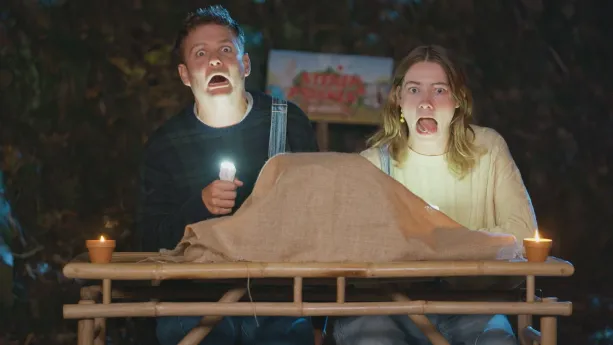 Een foto van presentatoren Jurre en Emma die achter een tafel staan waarop iets staat met een doek eroverheen. Ze hebben beide een zaklamp vast die ze op de onderkant van hun gezicht schijnen.