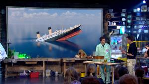 Een foto van presentator Jurre en Koen van Bemmelen in de studio achter een tafel met daarop een open aquarium dat half gevuld is met water en half met olie. Op de achtergrond is een scherm met daarop een animatie van het zinkende schip Titanic zichtbaar.