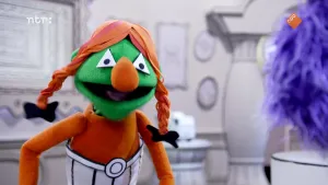 Een foto van een pop met oranjerode vlechten met zwarte strikjes erin en een witzwarte broek aan. De pop lijkt op Obelix uit de Asterix stripboeken. De pop heeft witte, driehoekige ogen, een grote oranje neus, een groen hoofd en oranjerode buik en armen.