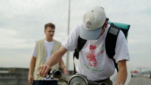 Een foto van presentator Sahil met een pet en rugtas op en heeft een shirt aan van een pizzabakker. Hij kijkt naar beneden terwijl hij op een scooter zit. Presentator Jurre staat op de achtergrond en kijkt toe.