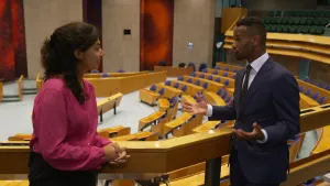 Een foto van presentator Manon die praat met Habtamu de Hoop. Op de achtergrond is de tweede kamer te zien met alle zetels.