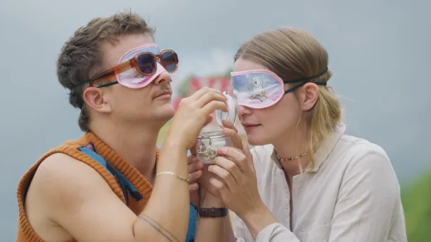 Een foto van presentatoren Emma en Jurre die met blinddoeken op een geopend potje vasthouden en erin ruiken.
