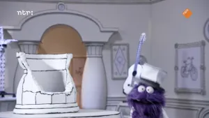 Een foto van een paarse pop in een zwart-wit getekende kamer. De pop heeft een hoed op en kijkt recht naar de kijker.