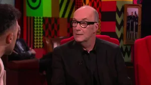 Een foto van een man die in de studio zit van Eus' Boekenclub. Links is het gezicht van de presentator zichtbaar, op de achtergrond zit er een man van het publiek.