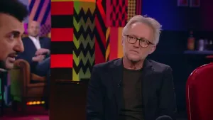 Een foto van een man die in de studio zit van Eus' Boekenclub. Links is het gezicht van de presentator zichtbaar, op de achtrond zit er een man in het publiek.