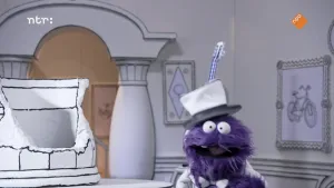 Een foto van een paarse pop in een zwart-wit getekende kamer. De pop heeft een hoed op en kijkt recht naar de kijker.