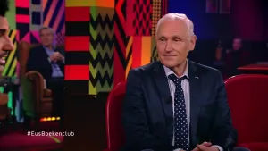 Een foto van een man die in de studio zit van Eus' Boekenclub. Links is het gezicht van de presentator zichtbaar, op de achtergrond zitten twee mensen van het publiek.