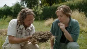 Een foto van presentator Pascal die samen met een vrouw op de grond zit met een hoop aarde in zijn handen. Ze zoeken naar insecten en de vrouw legt wat uit.