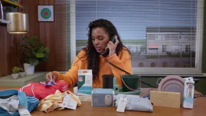 Een foto van een presentator die aan de telefoon zit te praten. Ze zit aan een tafel vol met producten die van plastic zijn gemaakt.